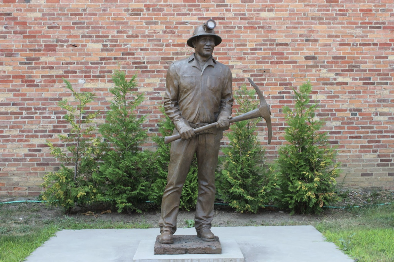 A bronze statue of a miner wearing coveralls, hard hat with light, and carrying a pickaxe. The statue stands in front of a brick building and small green shrubs.